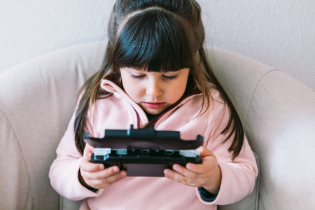 Little black haired girl wearing a pink sweater playing a video game with a portable video console...