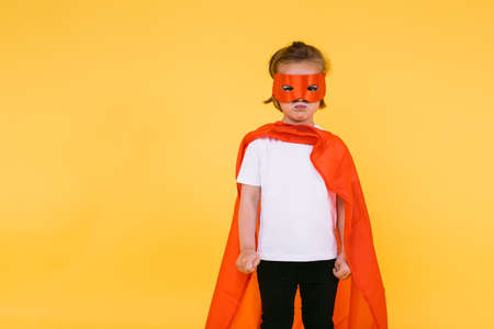 Little blonde girl dressed as a superheroine superhero with a cape and red mask, angry, with the cape to the side, on yellow backgroundの写真素材
