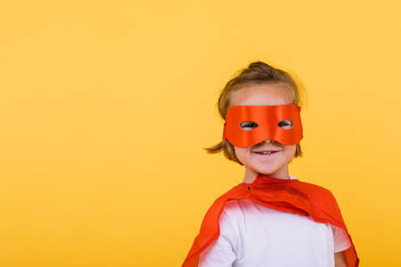 Little blonde girl dressed as superheroine superhero with cape and red mask, smiling, on yellow backgroundの写真素材