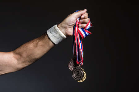 Arm of elderly athlete holding three gold, silver and bronze medals on a dark background. Sports and victory concept.の写真素材
