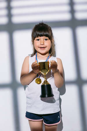 Dark-haired girl with sports championship medals and trophy cup, standing in the shadows of a window with a blind over her. Sport and victory conceptの写真素材