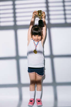 Dark-haired girl with sports championship medals, raising her trophy cup with her hands, over the shadows of a window with shutter above her. Sport and victory conceptの写真素材