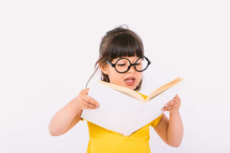 Smiling little girl wearing yellow t-shirt and round black glasses holding an open book in her hands and reading aloud on white backgroundの写真素材