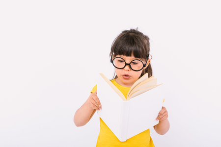 Surprised little girl, wearing yellow t-shirt and round black glasses holding an open book in her hands and reading, on white backgroundの写真素材