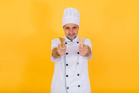 Chef cook wearing white kitchen cap and jacket with wooden spoon and fork, on yellow backgroundの写真素材