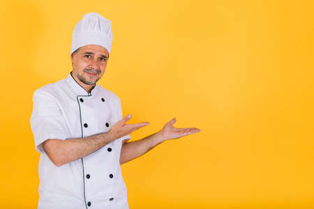 Chef cook wearing white kitchen cap and jacket pointing to the right with his hands, on yellow backgroundの写真素材