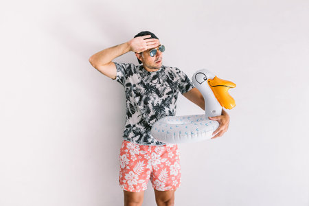 Young man with floral shirt, cap and glasses in summer with a duckling float making heat gestures,...