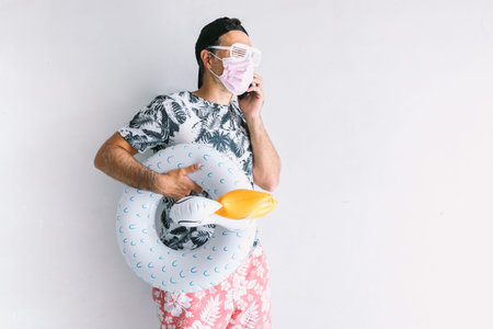 Young man wearing mask to protect himself from covid-19 on vacation, wearing a floral shirt and cap,...