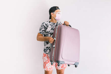 Young man wearing a mask to protect himself from covid-19 on vacation going on a trip, wearing a floral shirt and cap, holding a suitcase, in daylight on a white wallの写真素材