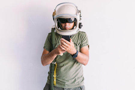Military man with astronaut cosmonaut helmet, looking at mobile phone, on a white wall
