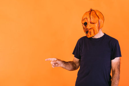 Man wearing terrifying pumpkin latex mask with blue t-shirt, pointing to the left side with his...