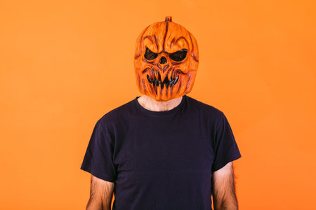 Man wearing scary pumpkin latex mask with blue t-shirt on orange background. Halloween and days of...