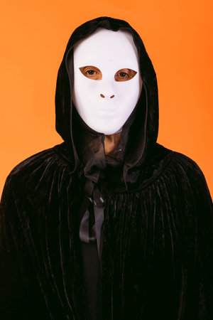 Portrait of a person in white killer Halloween mask, cape and hood looking at camera, dressed for...