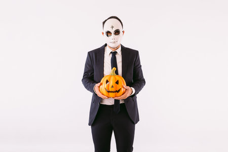 Man dressed in suit and tie, wearing a Halloween cross-browed killer mask, holding a pumpkin Jack-o-lantern. Carnival and halloween celebrationの写真素材