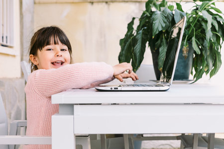 Side view of little girl wearing pink sweater, sitting in front of laptop, looking at camera, on terrace, smiling. Concept of distance studying online, childhood and technology.の写真素材