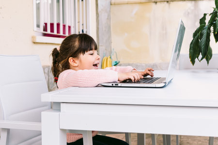 Side view of little girl wearing pink sweater, sitting in front of laptop, typing, on terrace, smiling. Concept of distance studying online, childhood and technology.の写真素材