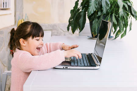 Side view of little girl wearing pink sweater, sitting in front of laptop, typing, on terrace, smiling. Concept of distance studying online, childhood and technology.の写真素材