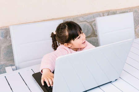 Front view of little girl wearing pink sweater, sitting in front of laptop, on terrace, smiling. Concept of distance studying online, childhood and technology.の写真素材