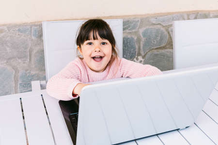 Front view of little girl wearing pink sweater, sitting in front of laptop, looking at camera, on terrace, smiling. Concept of distance studying online, childhood and technology.の写真素材