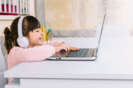 Little girl with white headphones, wearing pink sweater, sitting in front of laptop, typing, on terrace, smiling. Concept of distance studying online, childhood and technology.の写真素材