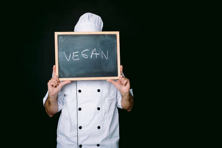 Cook dressed in a white jacket and hat holds a blackboard that reads: "Vegan" on a black background. Vegan food and restaurant concept.の写真素材