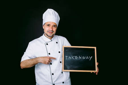 Cook dressed in a white jacket and hat holding a blackboard that reads: "Take away" on a black background. Restaurant and takeaway concept.の写真素材