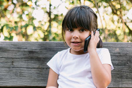 Little black-haired girl, wearing a white t-shirt, sitting on a park bench talking on the mobile phone, smiling.の写真素材