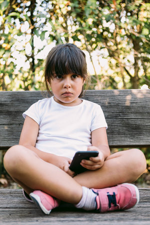 Little girl with black hair, wearing a white T-shirt, sitting on a bench in a park, looking at the...