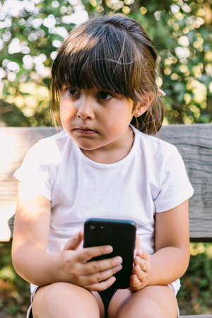 Little girl with black hair, wearing a white T-shirt, sitting on a bench in a park, looking at the mobile phone, with a serious gesture.の写真素材