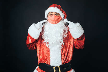 Man dressed as Santa Claus with a red mask, pointing at it with his fingers, on black background. Concept of Christmas, Santa Claus, gifts, celebration.の写真素材