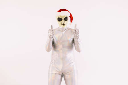Person dressed in silver suit and green alien mask, with a Santa Claus Christmas hat, raising thumbs up, on white backgroundの写真素材