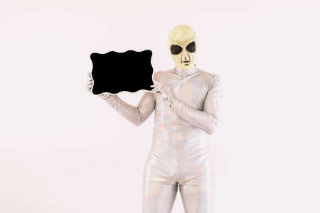 Person dressed in silver suit and green alien mask, holding a blackboard, on white backgroundの写真素材