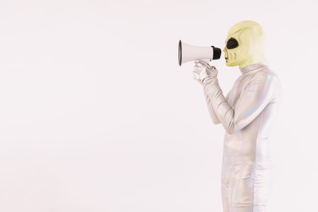 Person dressed in silver suit and green alien mask, speaking with a white megaphone, on white...