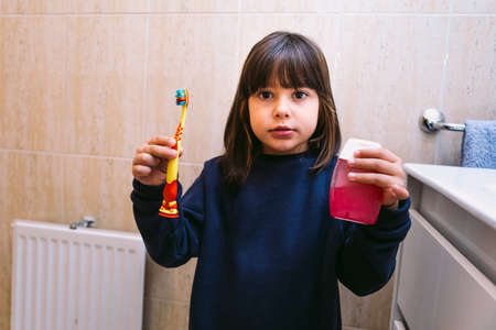 Small girl in dark blue sweatshirt, holding brush her toothbrush and toothpaste, in bathroom. Tooth brushing, hygiene and childhood concept.の写真素材