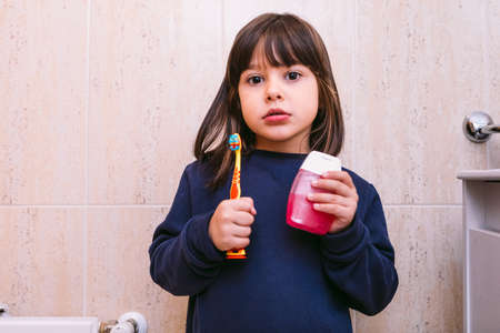 Small girl in dark blue sweatshirt, holding brush her toothbrush and toothpaste, in bathroom. Tooth brushing, hygiene and childhood concept.の写真素材