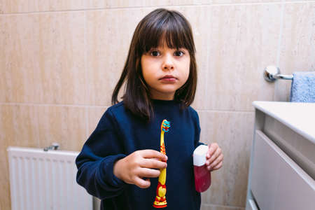 Small girl in dark blue sweatshirt, holding brush her toothbrush and toothpaste, in bathroom. Tooth brushing, hygiene and childhood concept.の写真素材