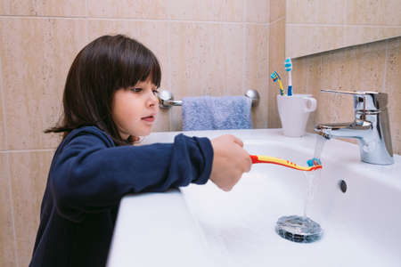 Little girl in a dark blue sweatshirt, brushing her teeth and pouring water on the toothbrush, in the bathroom. Tooth brushing, hygiene and childhood concept.の写真素材