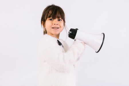 Little black haired girl wearing white sweater with bow tie, speaking through a white megaphone.の写真素材
