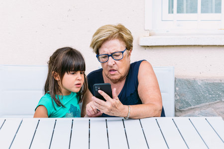Grandmother and granddaughter with a strange face consulting the mobile on a terrace. Family,...