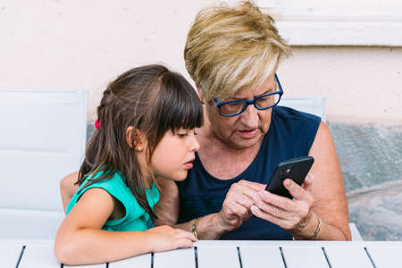 Grandmother and granddaughter consulting the mobile on a terrace. Family, grandchildren, grandparents and technology concept.の写真素材