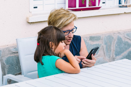 Happy grandmother and granddaughter consulting the mobile on a terrace. Family, grandchildren, grandparents and technology concept.の写真素材