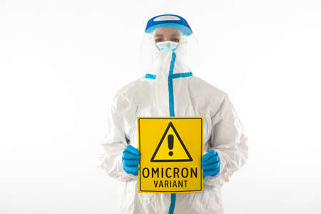 Medical nurse wearing a PPE and latex gloves holding a yellow sign with a danger symbol that says: 'VARIANT OMICRON'. Coronavirus, pandemic and health concept.の写真素材