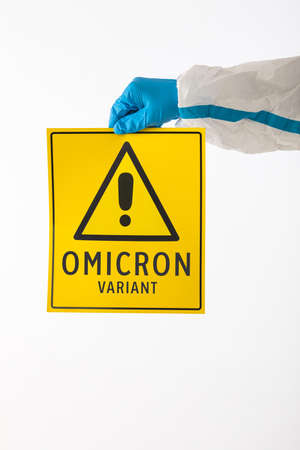 Detail of an arm of a medical nurse wearing a PPE and latex gloves with a yellow sign with a danger symbol that reads: 'VARIANT OMICRON'. Coronavirus, pandemic and health concept.の写真素材