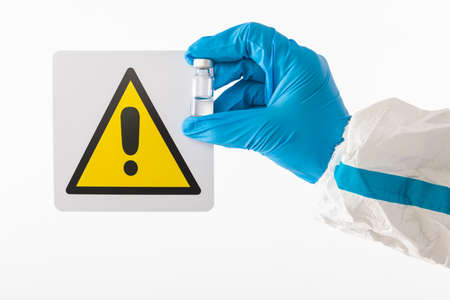 Details of the arms of a medical nurse wearing PPE and latex gloves holding a sign with the danger symbol and a vaccination vial with the vaccine inside. Coronavirus, pandemic, omicron variant and health concept.の写真素材