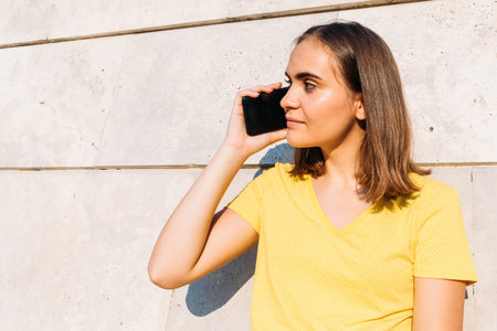 Young blond-haired girl wearing yellow t-shirt, talking with her smart phone leaning against a stone...