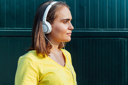 Young blond-haired girl wearing yellow t-shirt, listening to music with white headphones, on green...