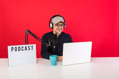 Male presenter with cap in podcast radio recording studio smiling and consulting the Internet, next to a computer, a microphone and a light box with the word podcast, on a red background.の写真素材