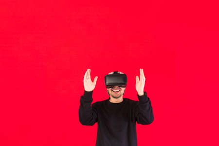 Man with cap dressed in black with Virtual Reality glasses smiling while touching something virtually, on red background, with copy space. Technology, virtual reality, computing and hobbies concept.の写真素材