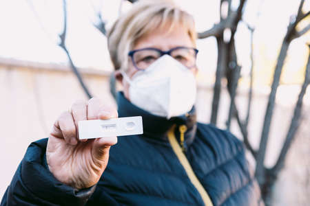 Old lady in the street, with a white mask, out of focus, holding a negative antigen test. Concept covid-19, ihu, delta, omicron, detection test and pandemic.の写真素材