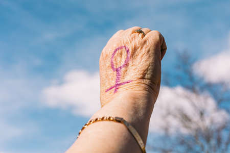 Fist of an older woman with purple painted nails, with the sky in the background, with the female symbol painted. Concept of women's day, empowerment, equality, inequality, activism and protest.の写真素材
